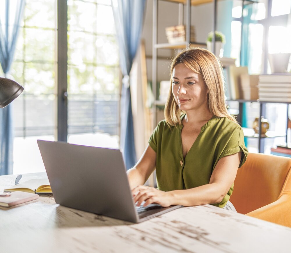 Eine Frau mit hellbraunem Haar und grüner Bluse sitzt an einem Holzschreibtisch und arbeitet an einem Laptop in einem hellen, modernen Heimbüro mit großen Fenstern und Regalen im Hintergrund.