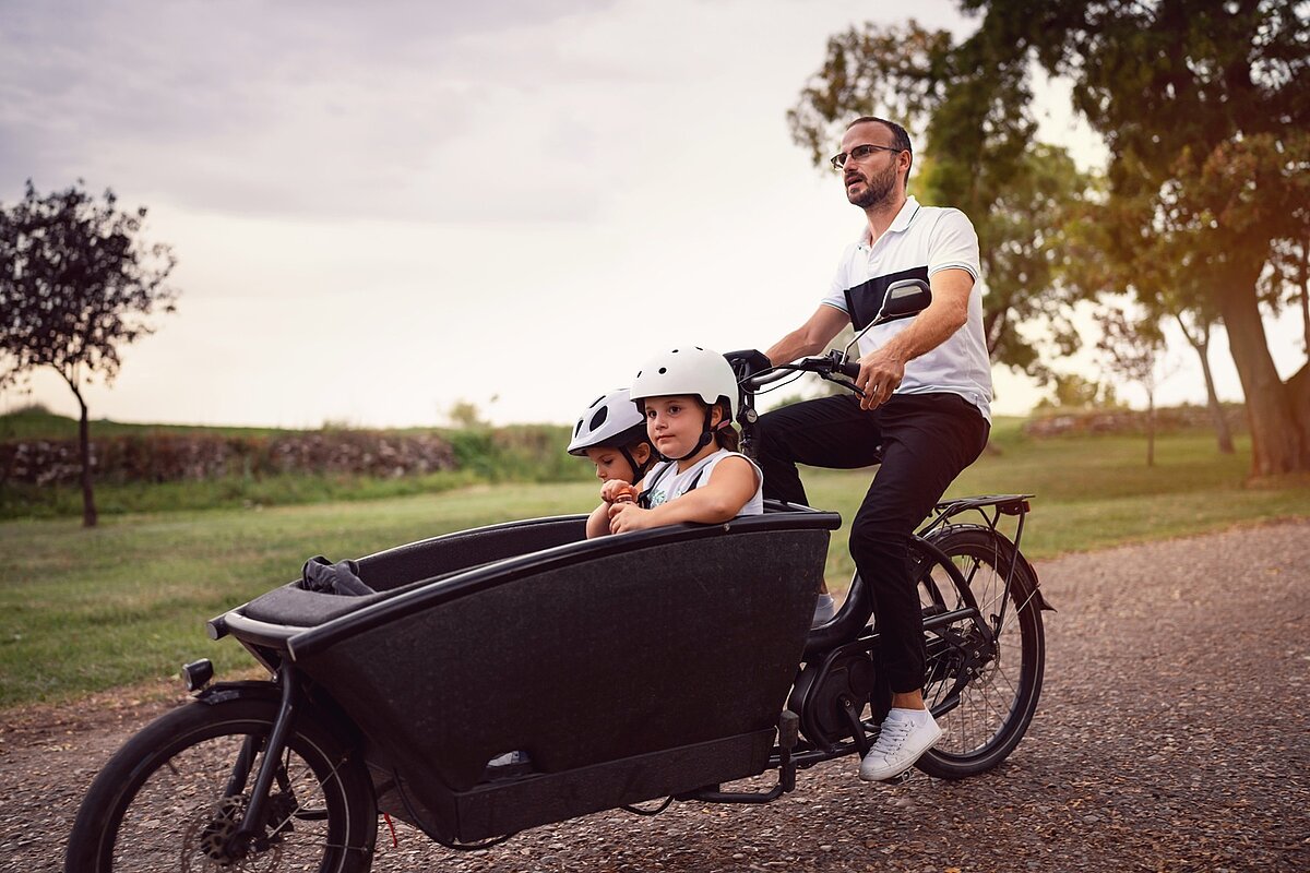 Vater transportiert Kind im Lastenrad mit Frontbox auf einem Feldweg.