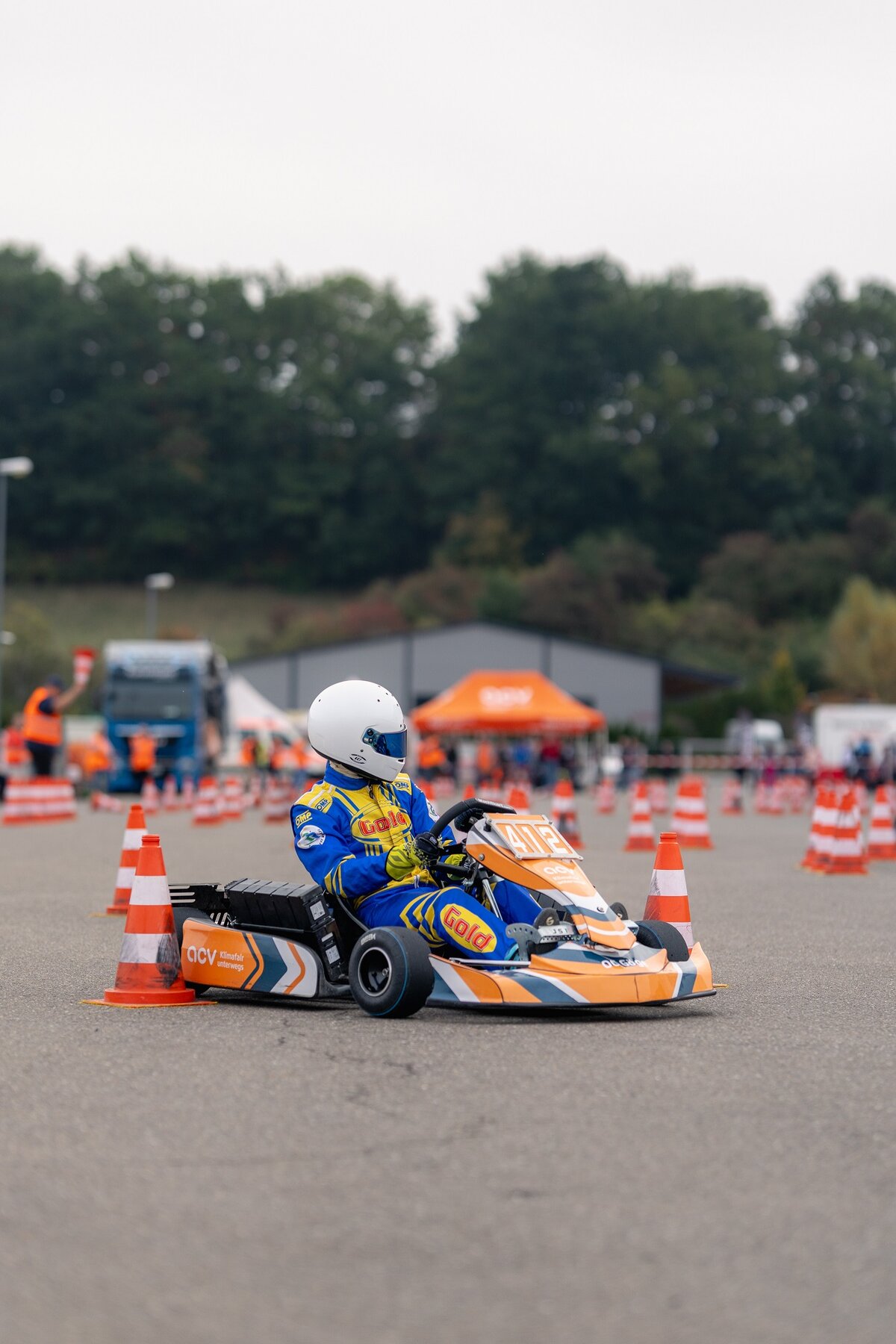 Ein Go-Kart-Fahrer in einem blau-gelben Rennanzug und einem weißen Helm schlängelt sich durch einen Parcours aus orangefarbenen Verkehrskegeln.