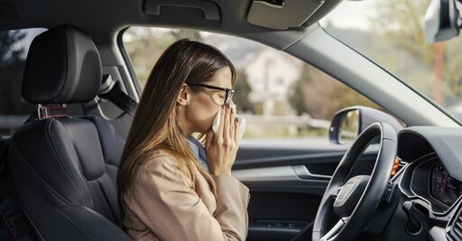 Eine Frau sitzt auf dem Fahrersitz eines Autos, trägt eine Brille und eine beigefarbene Jacke und hält sich ein Taschentuch vor die Nase, als ob sie niesen müsste oder sich unwohl fühlt. Das Auto ist geparkt und der Innenraum ist sichtbar.