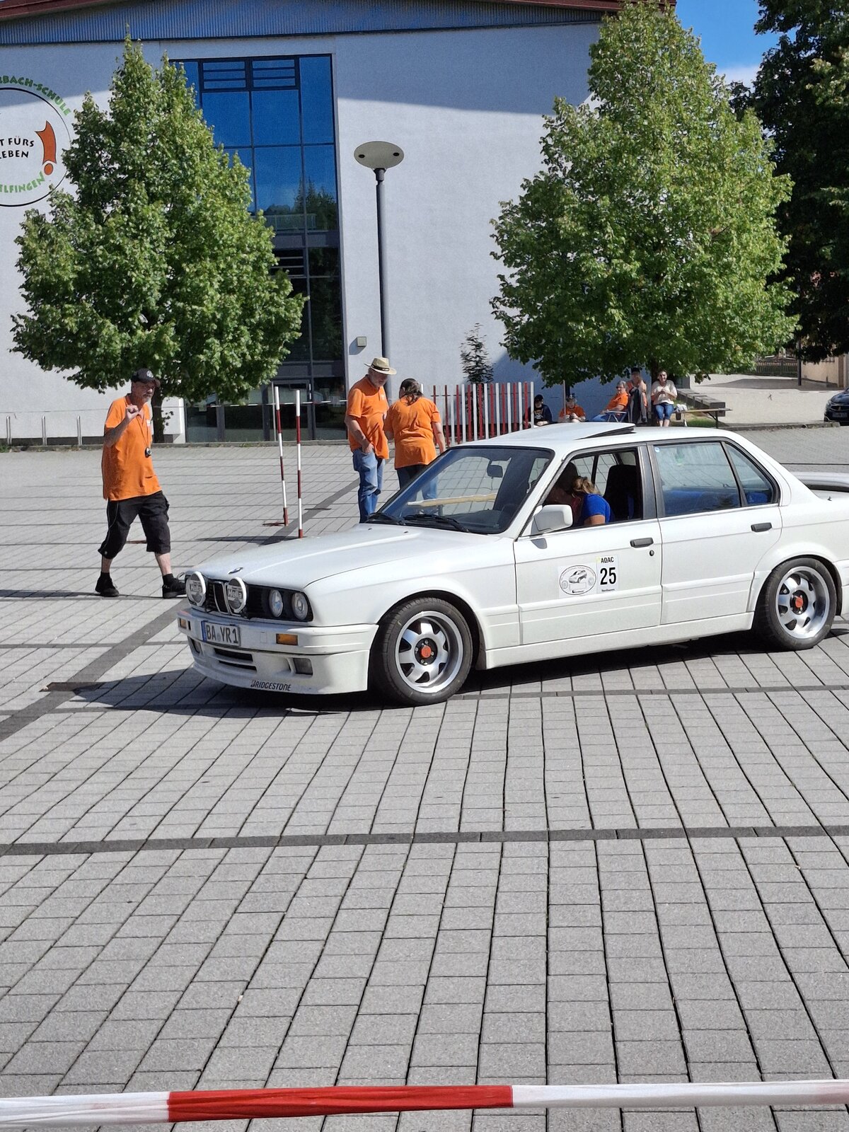 Ein weißer BMW mit der Nummer 25 an der Seite fährt auf einer asphaltierten Fläche, während im Hintergrund mehrere Personen in orangefarbenen Hemden in der Nähe einiger Bäume und eines weißen Gebäudes stehen.