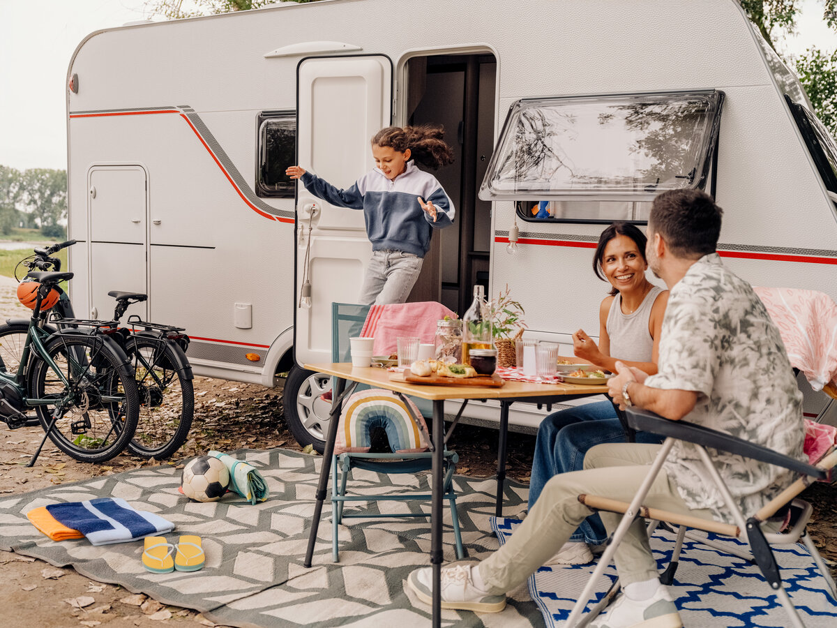 „Familie beim Camping sitzt vor einem Wohnwagen am Tisch, während ein Kind aus der Tür des Wohnwagens springt.“
