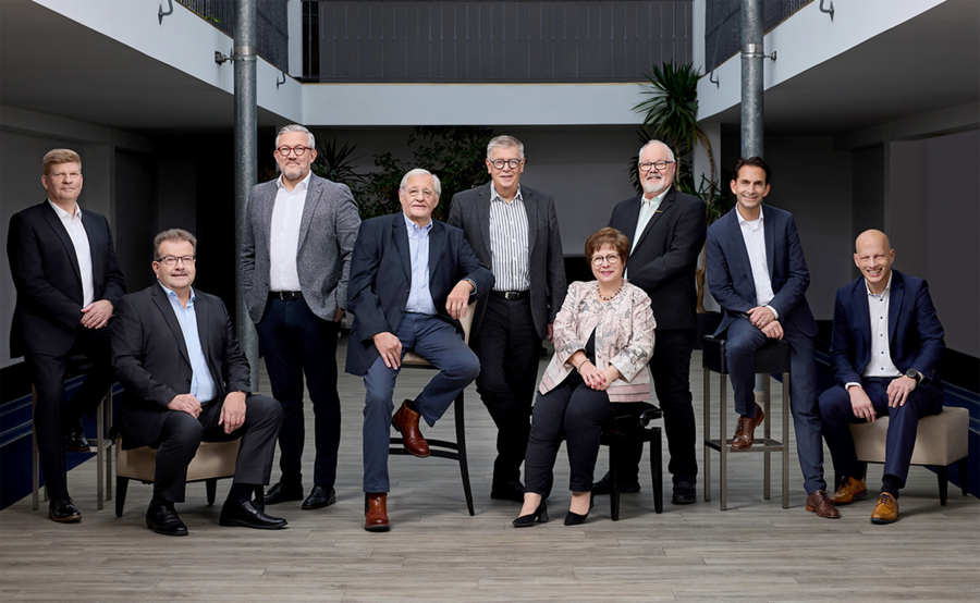 Neun professionell gekleidete Personen, acht Männer und eine Frau, posieren gemeinsam in einem Innenraum. Die meisten stehen, während drei auf Stühlen oder Hockern sitzen, in einem modernen Büroraum mit Pflanzen und einem Balkon im Hintergrund.