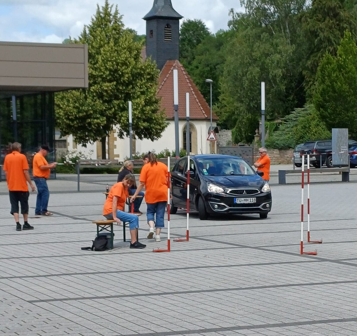 Mehrere Personen in orangefarbenen Hemden richten auf einer gepflasterten Fläche mit Stangen eine Veranstaltung im Freien ein. In der Nähe hält ein schwarzes Auto, und im Hintergrund sind Bäume und Gebäude, darunter eine Kirche, zu sehen.