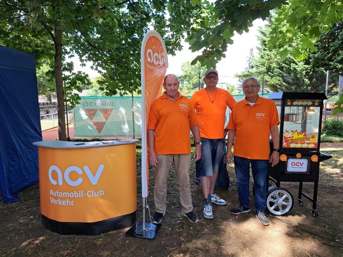 Drei Männer in orangefarbenen ACV-T-Shirts stehen an einem Stand im Freien unter Bäumen, neben einem orangefarbenen ACV-Tisch und einem Popcorn-Wagen, und werben für den Automobil-Club Verkehr. Im Hintergrund sind der Zaun eines Sportplatzes und ein Banner zu sehen.