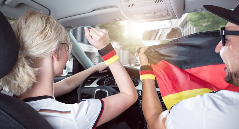 Zwei Personen in einem Auto, die weiße Hemden und Armbänder mit den Farben der deutschen Flagge tragen, feiern, während sie eine große deutsche Flagge halten. Der Fahrer hebt begeistert eine Faust. Das Sonnenlicht scheint durch die Windschutzscheibe.