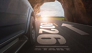 Blick aus einem dunklen Tunnel auf eine helle Straße mit der auf den Asphalt gemalten Aufschrift „2026“, die in Richtung Tageslicht und offener Landschaft führt; links ist ein Teil eines Fahrzeugs sichtbar.