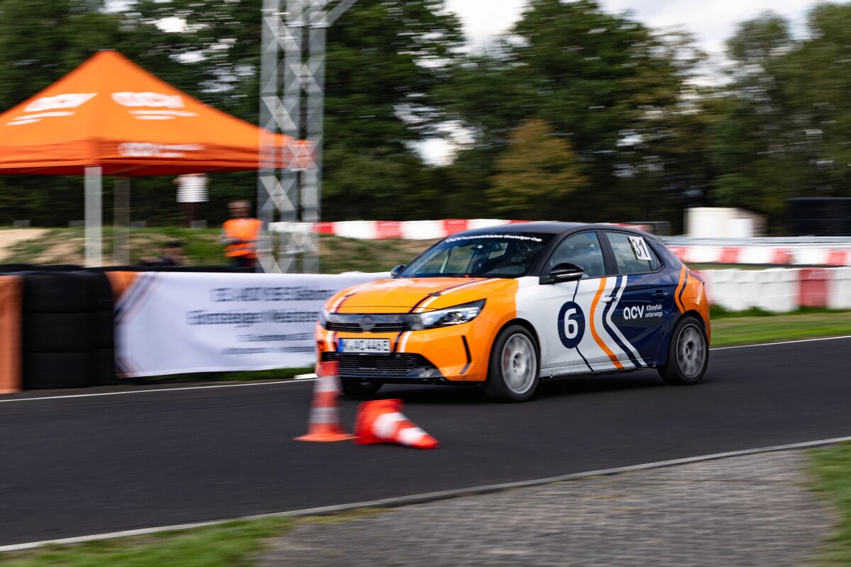 Ein orange-weißer Rennwagen mit der Nummer 6 rast auf einer Rennstrecke in der Nähe von orangefarbenen Kegeln und einem Zelt vorbei, während im Hintergrund eine Person in einer orangefarbenen Weste steht. Hinter den Absperrungen sind Bäume und ein Transparent zu sehen.