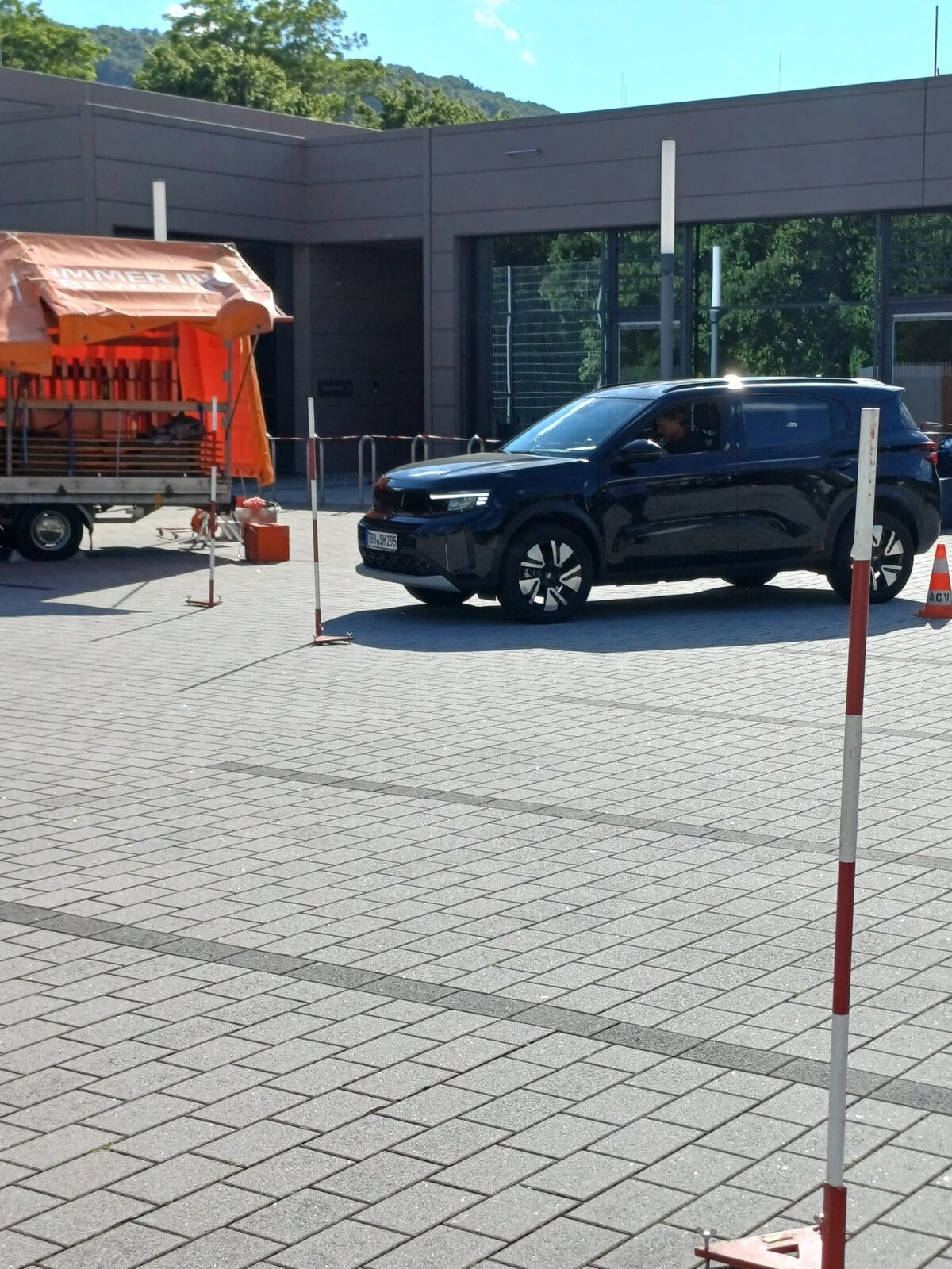 Ein schwarzer Geländewagen fährt auf einer asphaltierten Fläche in der Nähe einer Baustelle mit orangefarbenen Absperrungen und Leitkegeln. Links ist ein Anhänger mit Baumaterialien zu sehen, und die Szene wird von einem modernen Gebäude eingerahmt.