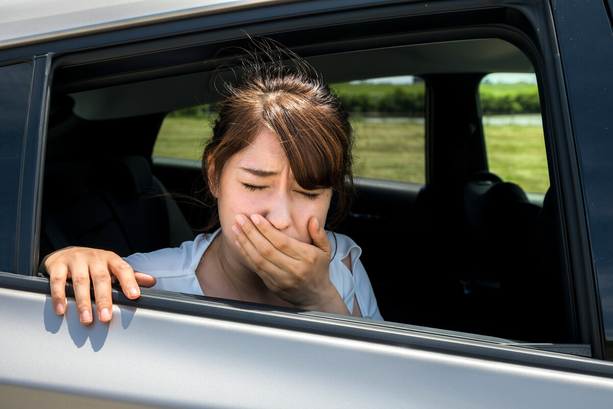 Eine Frau lehnt sich aus einem Autofenster, hält sich den Mund mit der Hand zu und scheint sich unwohl zu fühlen, möglicherweise leidet sie an Reisekrankheit. Außerhalb des Autos sind Bäume und Gras zu sehen.