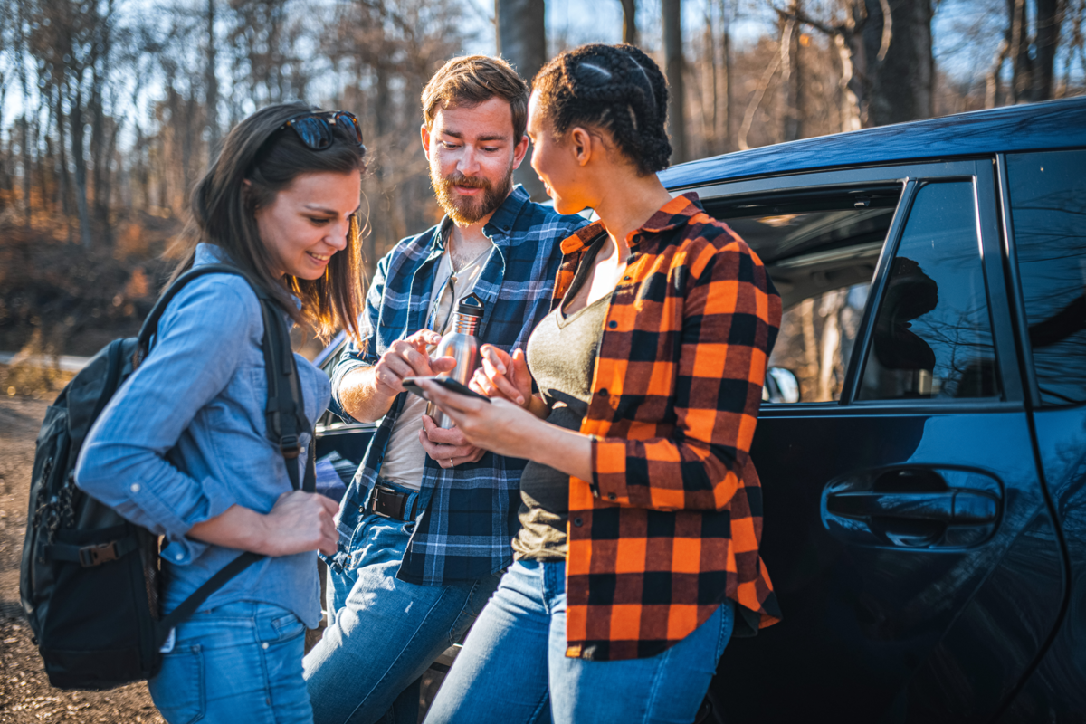 2 junge Frauen und ein junger Mann stehen angelehnt an ein Auto in einem Wald und zeigen auf ein Smartphone