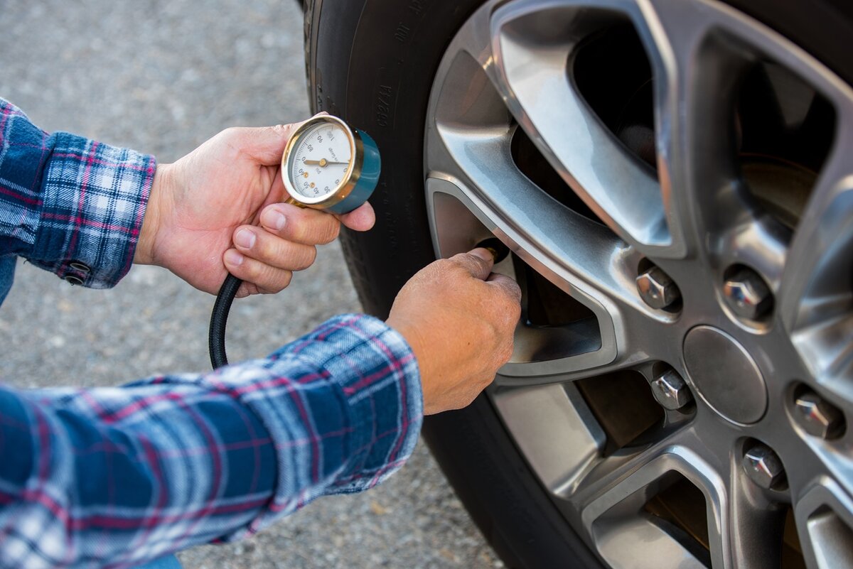 Eine Person in einem karierten Hemd überprüft mit einem Reifendruckmesser den Luftdruck in einem Autoreifen auf einer asphaltierten Fläche.