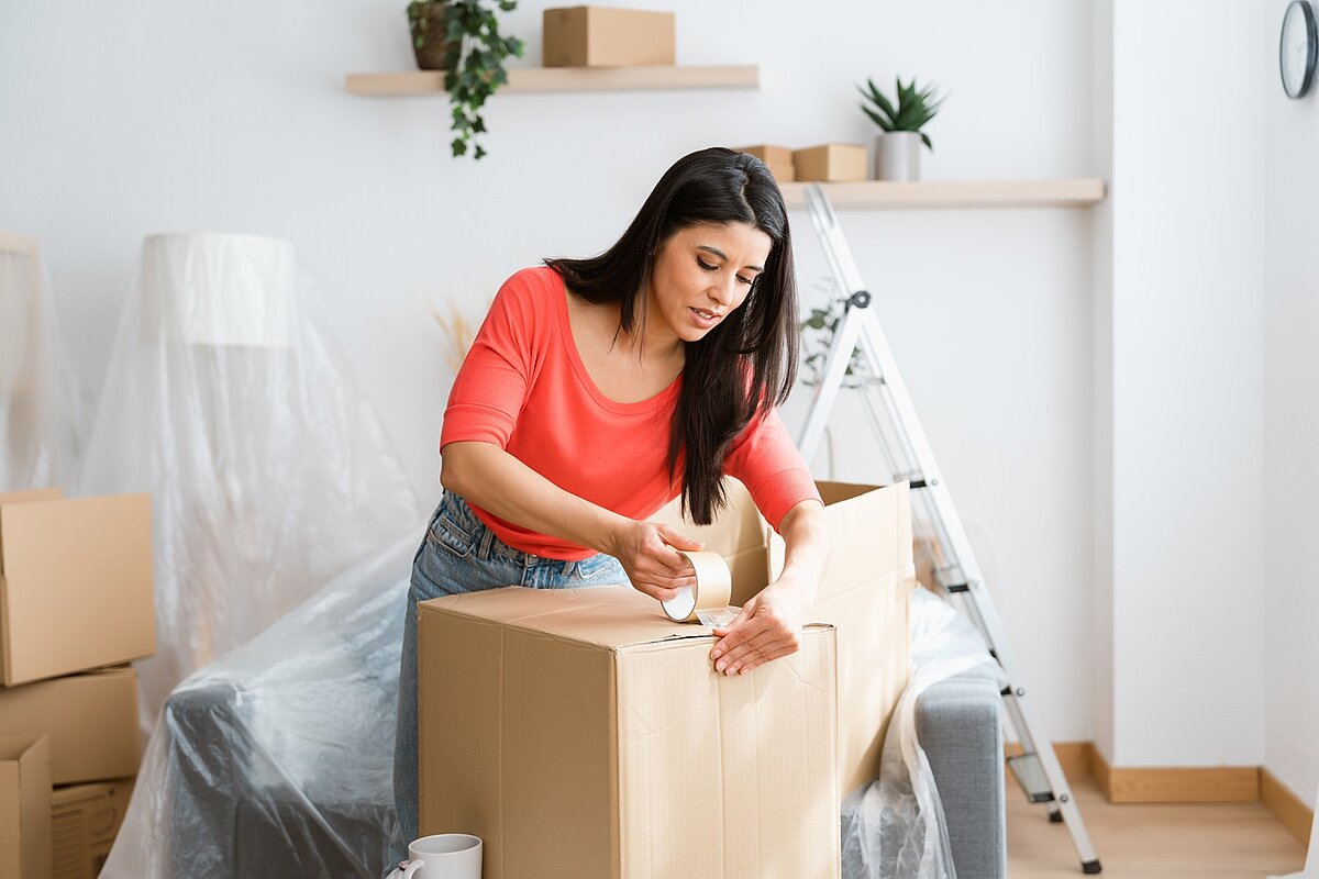 Eine Frau steht in einem hellen Raum und verschließt konzentriert einen Umzugskarton. Hinter ihr stehen verpackte Möbel, Kartons und eine Leiter.