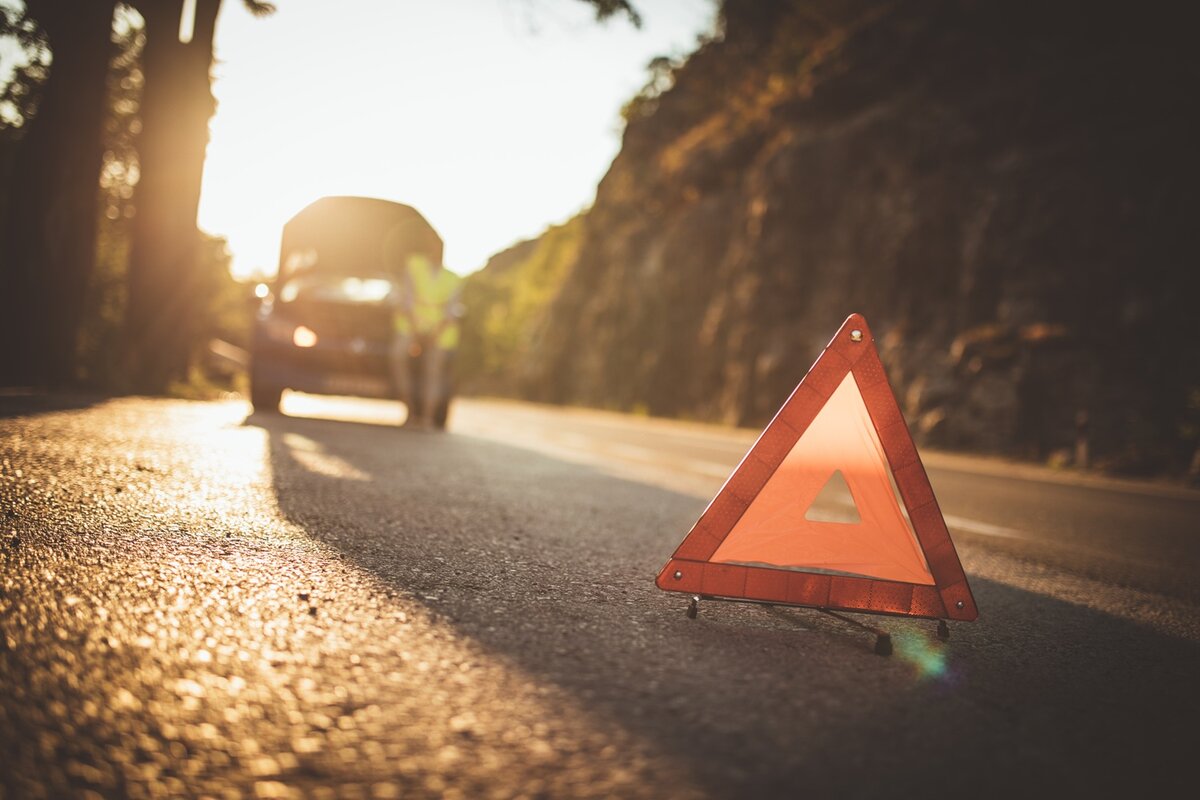 Warndreieck neben einem liegen gebliebenen Auto auf einer Landstraße bei tief stehender Sonne.