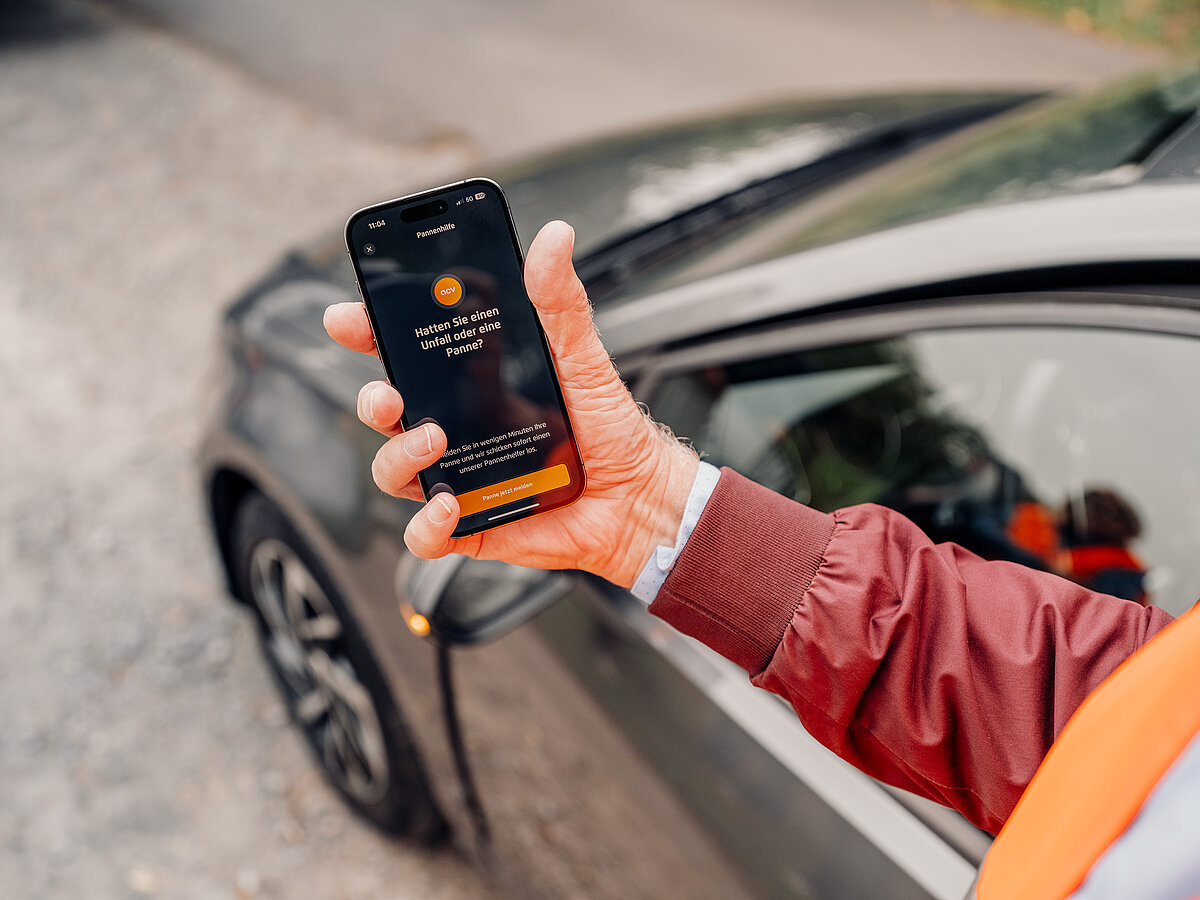 „Person in Warnweste hält ein Smartphone mit geöffneter ACV-App neben einem Auto.“