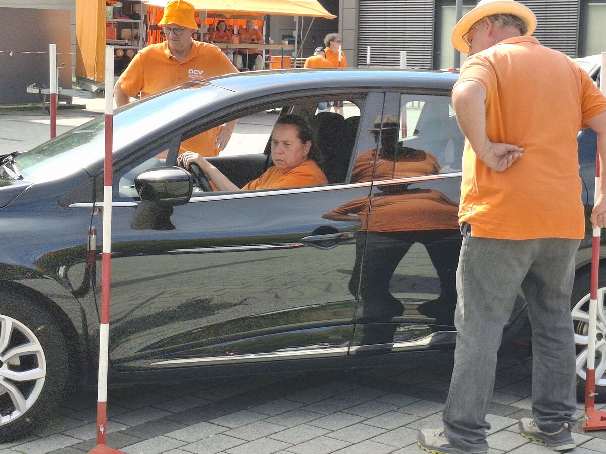 Eine Frau sitzt auf dem Fahrersitz eines schwarzen Autos und blickt zu einem Mann in einem orangefarbenen Hemd und einer orangefarbenen Mütze, der draußen steht. Im Hintergrund sind zwei weitere Personen in orangefarbenen Hemden zu sehen, die neben orangefarbenen Geräten stehen.