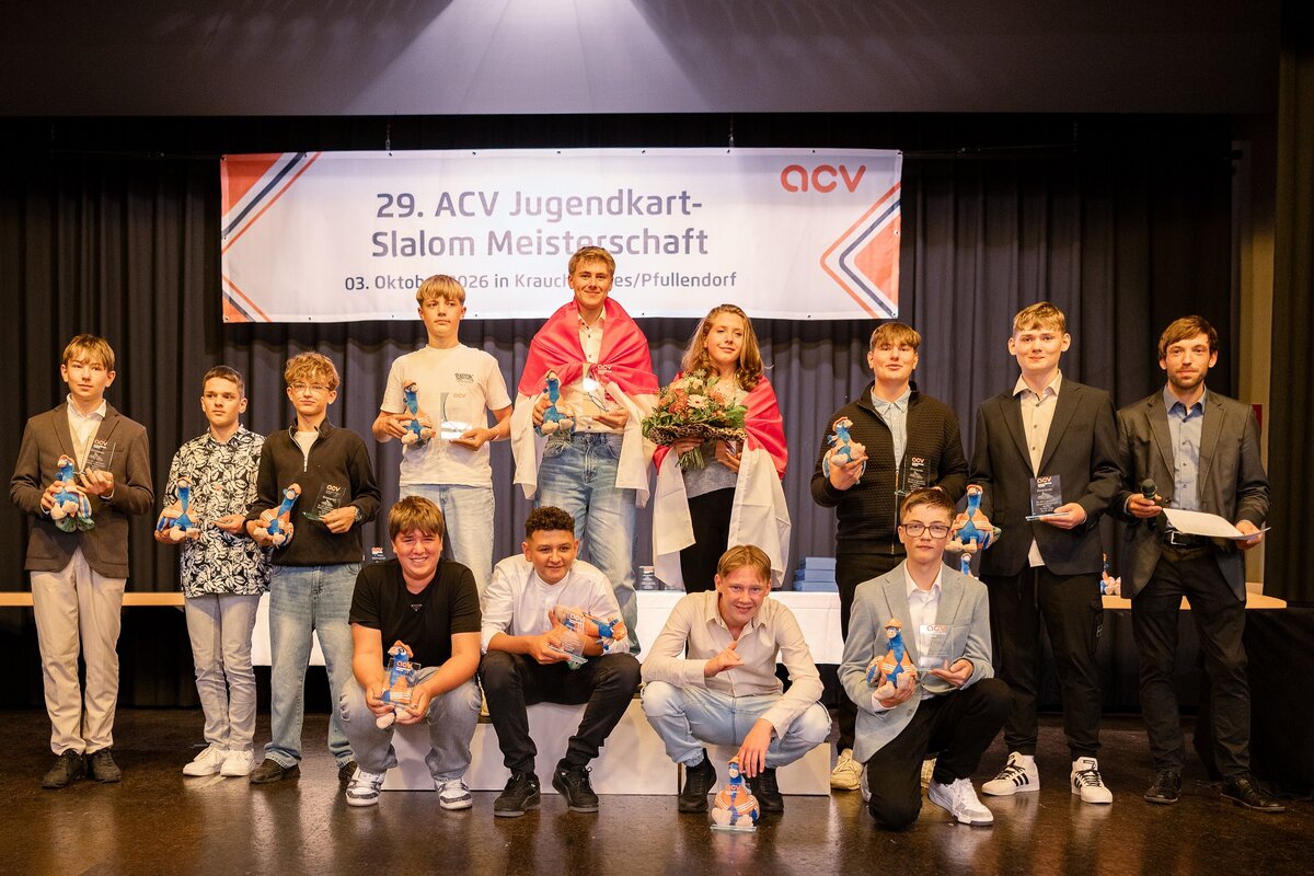 Eine Gruppe junger Menschen posiert bei der Siegerehrung der 29. ACV-Jugendkart-Slalom-Meisterschaft auf der Bühne für ein Foto und hält dabei Pokale und kleine blaue Figuren in den Händen.