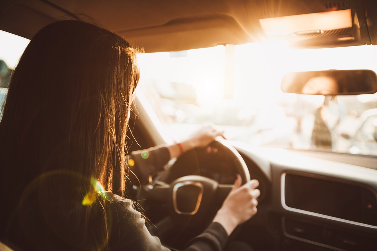 Eine Person mit langen Haaren fährt bei Sonnenuntergang mit dem Auto; das Sonnenlicht strömt durch das Fenster und spiegelt sich auf dem Armaturenbrett wider, wodurch im Innenraum ein warmer Schein entsteht.