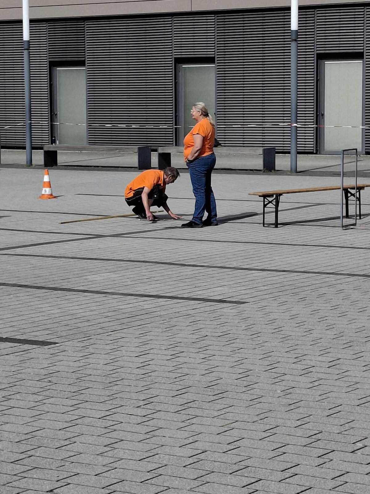 Zwei Personen in orangefarbenen Hemden arbeiten im Freien auf einer gepflasterten Fläche; eine kniet neben einem Maßband auf dem Boden, während die andere neben einer Bank steht. Im Hintergrund sind ein Verkehrskegel und Gebäudetüren zu sehen.