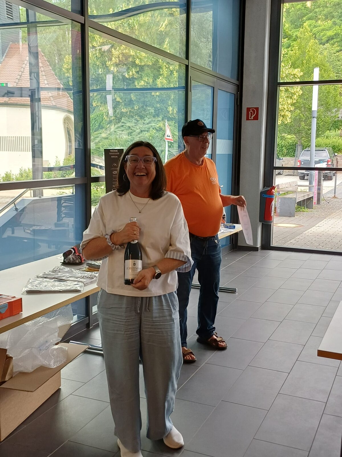 Eine lächelnde Frau in einem weißen Oberteil und einer grauen Hose hält in einem modernen Gebäude mit Glasfassade eine Weinflasche in der Hand. Hinter ihr steht ein Mann in einem orangefarbenen Hemd in der Nähe des Eingangs.