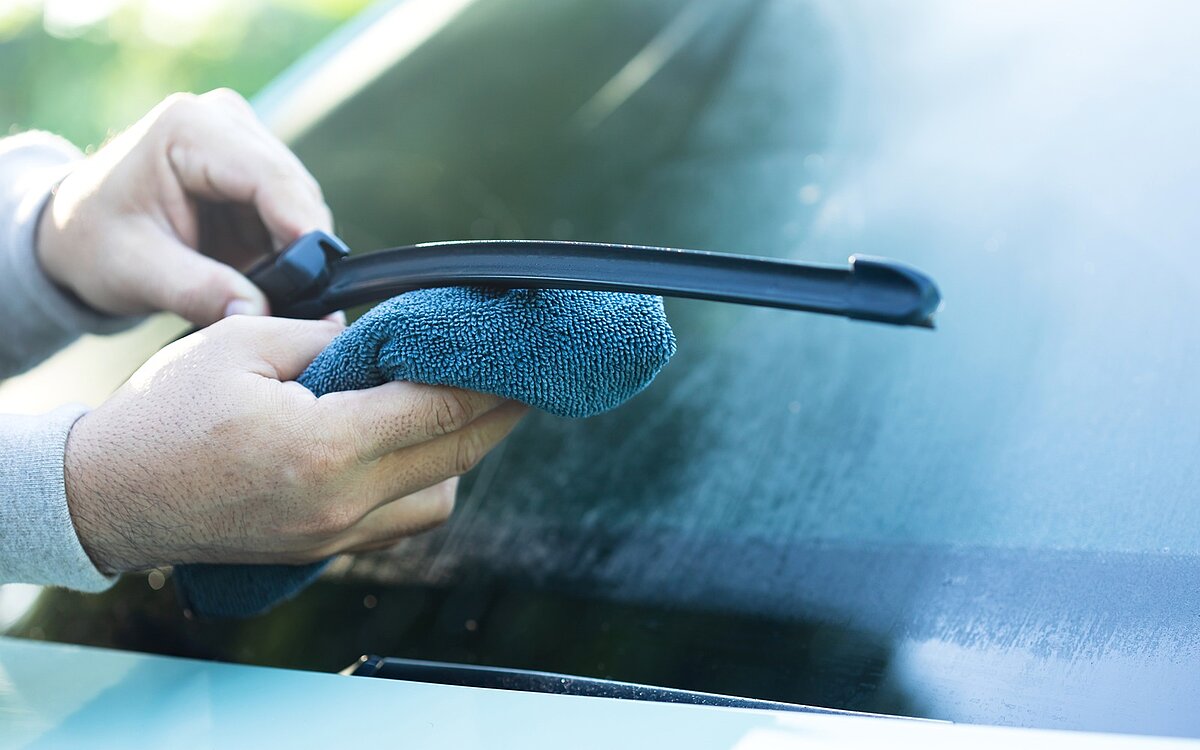 Nahaufnahme einer Person, die ein Scheibenwischerblatt mit einem blauen Mikrofasertuch reinigt, während der Wischer von der Windschutzscheibe des Autos abgehoben wird.