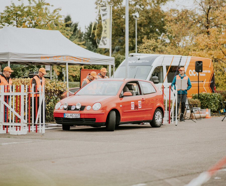 Ein kleines rotes Auto wartet an einem Kontrollpunkt, der von Personen mit orangefarbenen Sicherheitswesten und Mützen besetzt ist. Im Hintergrund sind ein weißes Vordach und ein orangefarbener Lieferwagen zu sehen, das Gelände ist von Bäumen und Zäunen umgeben.