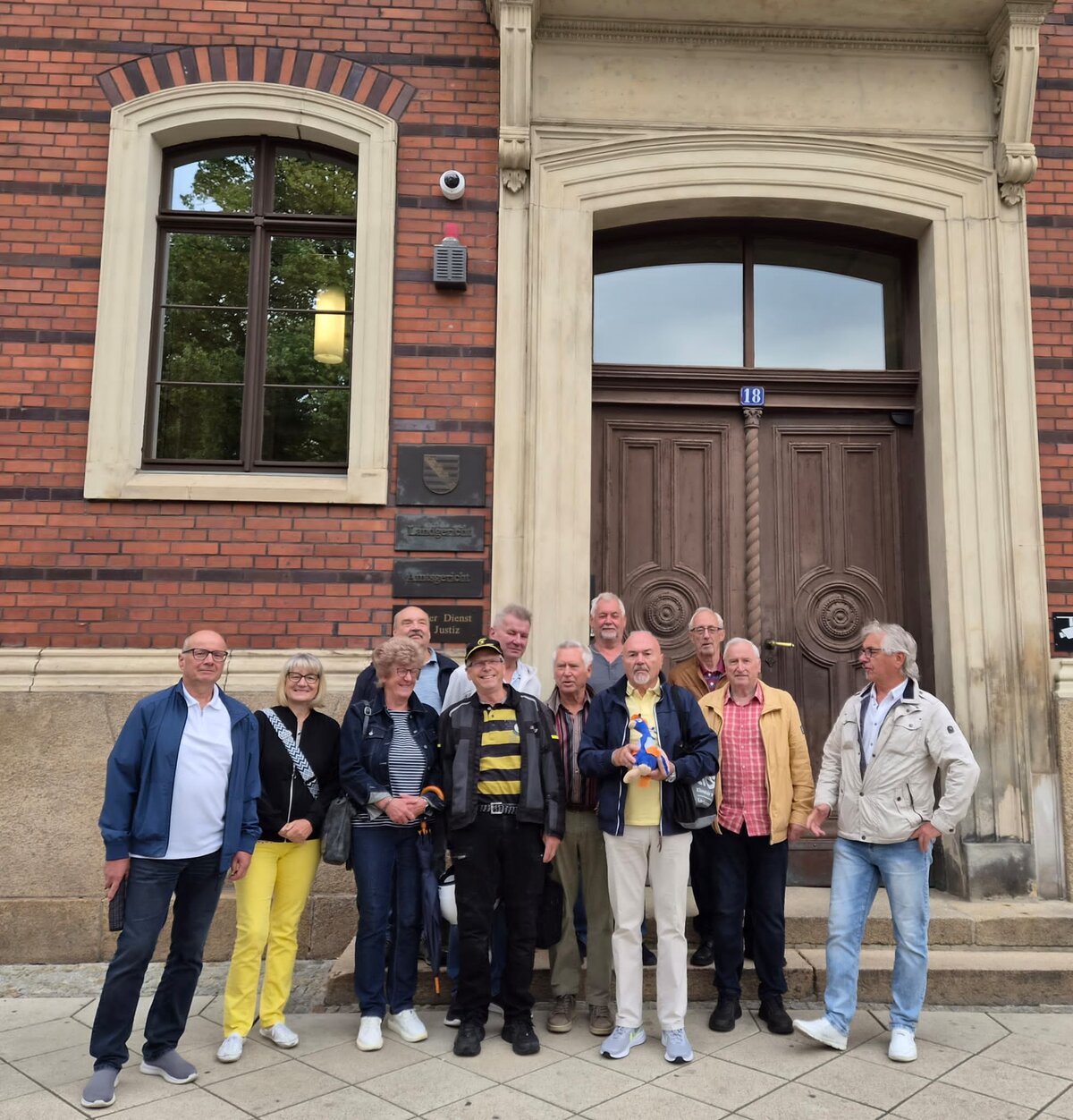 Eine Gruppe von zwölf Erwachsenen steht gemeinsam vor einem Backsteingebäude mit einer großen Holztür und einem gewölbten Steinrahmen und posiert auf dem Bürgersteig für ein Gruppenfoto.