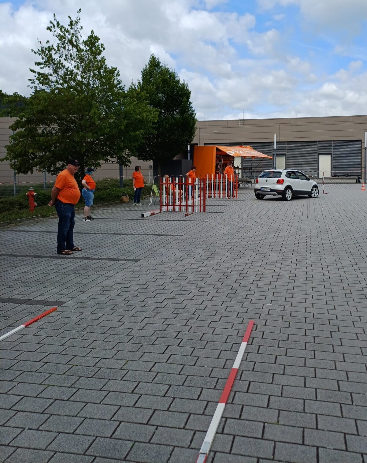 Menschen in orangefarbenen Hemden stehen neben einem mobilen Kiosk, roten und weißen Stangen und einem weißen Auto, das auf einem gepflasterten Parkplatz unter bewölktem Himmel geparkt ist.