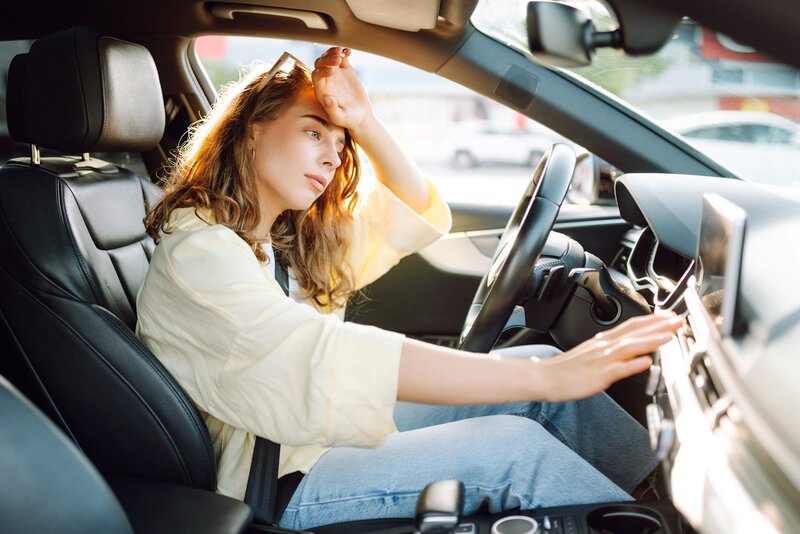Eine Frau sitzt in einem Auto, wirkt müde und erhitzt und streckt die andere Hand nach der Klimaanlage aus.