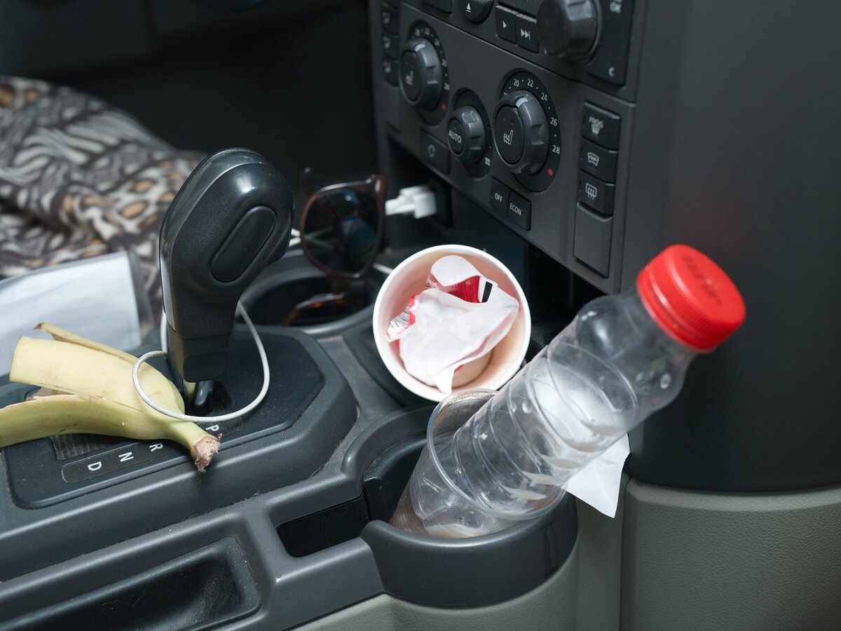 Die Mittelkonsole eines Autos mit einer Bananenschale, einer Sonnenbrille, einer leeren Wasserflasche und einem mit Müll gefüllten Becher – ein Bild, das einen unaufgeräumten und chaotischen Innenraum zeigt.