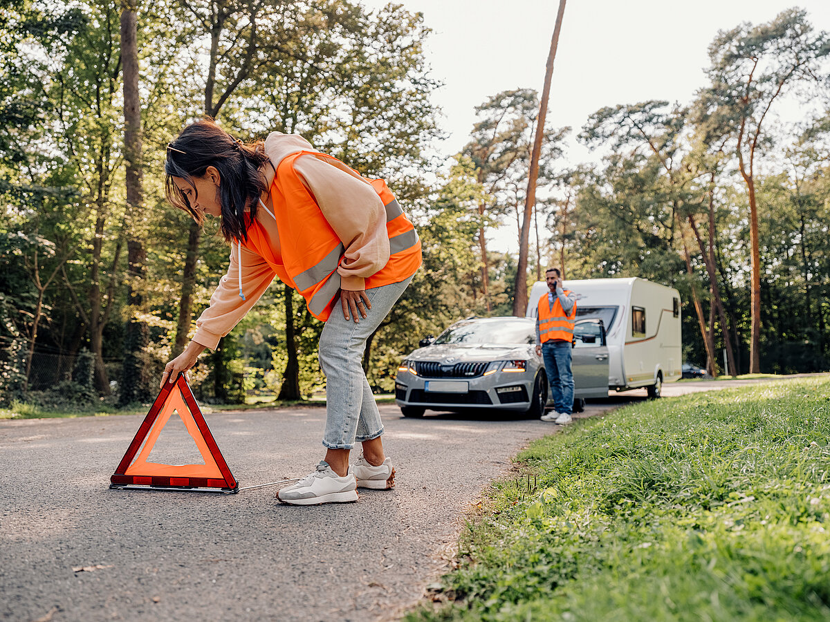 „Frau in Warnweste stellt ein Warndreieck auf die Straße, im Hintergrund stehen ein Auto mit Wohnwagen und eine weitere Person in Warnweste.“