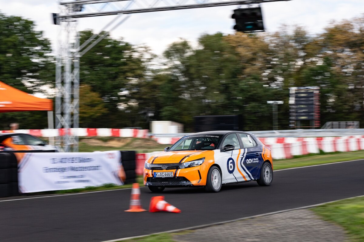 Ein orange-weißer Rennwagen mit der Startnummer 6 rast auf einer Rennstrecke an orangefarbenen Kegeln und einem Banner vorbei. Im Hintergrund sind Bäume und Absperrungen zu sehen, und eine weitere Person steht in der Nähe eines orangefarbenen Zeltes.