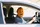 	Eine Person sitzt am Steuer eines Autos und trägt eine große Kürbis-Maske mit geschnitztem Gesicht. Sie trägt ein kariertes Hemd und hält das Lenkrad mit beiden Händen.