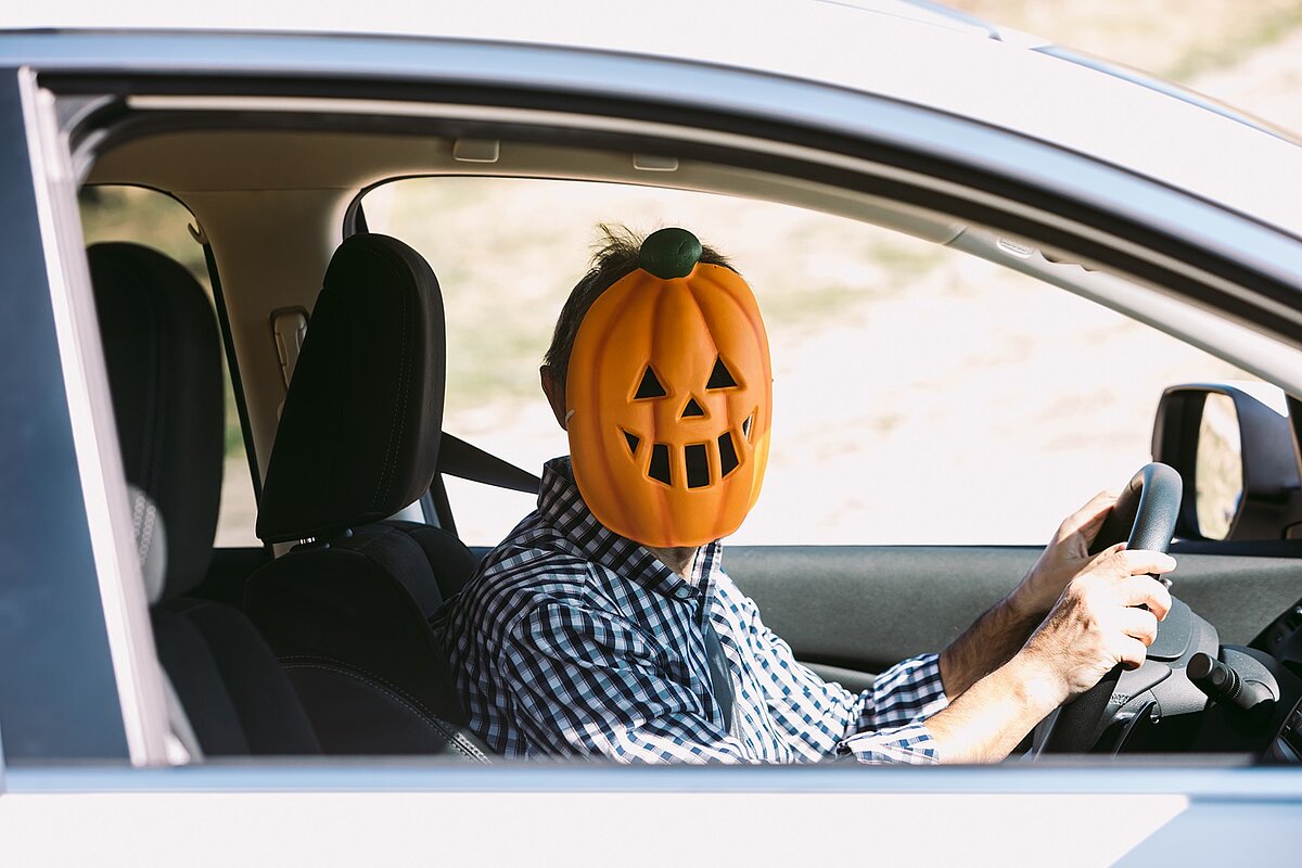 	Eine Person sitzt am Steuer eines Autos und trägt eine große Kürbis-Maske mit geschnitztem Gesicht. Sie trägt ein kariertes Hemd und hält das Lenkrad mit beiden Händen.