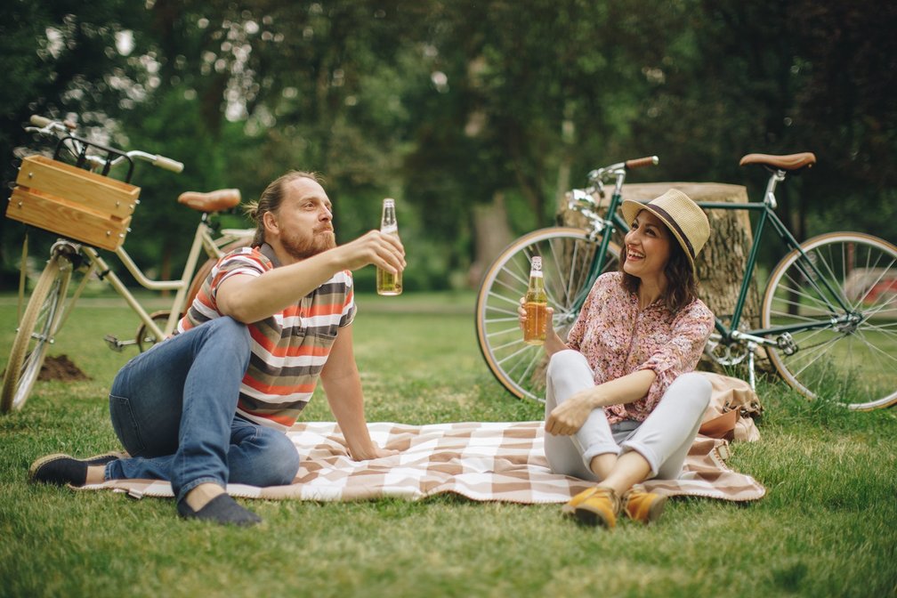 Zwei Menschen sitzen auf einer Picknickdecke im Park und trinken Bier, im Hintergrund stehen zwei Fahrräder.
