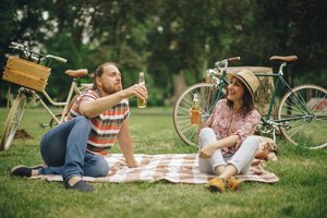 Zwei Menschen sitzen auf einer Picknickdecke im Park und trinken Bier, im Hintergrund stehen zwei Fahrräder.