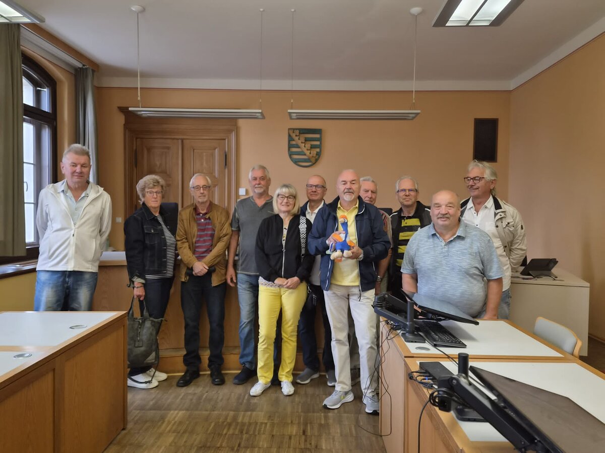 Elf Erwachsene stehen in einem Klassenzimmer oder Besprechungsraum mit Holzboden und lächeln gemeinsam. An der Wand hinter ihnen ist ein Wappen zu sehen, im Vordergrund stehen Schreibtische mit Computerbildschirmen.