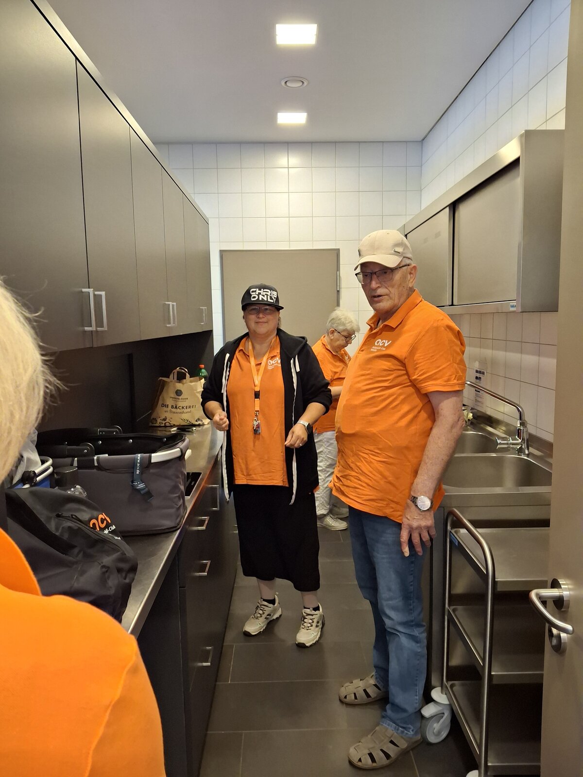Vier Erwachsene in orangefarbenen Hemden und Mützen stehen in einer modernen Küche mit grauen Schränken und gefliesten Wänden. Zwei davon befinden sich im Vordergrund, einer steht in der Nähe der Schränke und ein weiterer ist im Hintergrund zu sehen, wo er teilweise verdeckt ist.