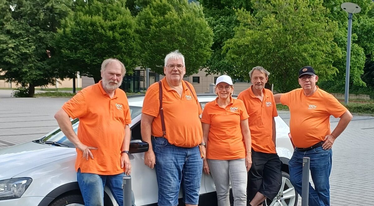 Fünf Personen in orangefarbenen T-Shirts stehen neben einem weißen Auto. Sie lächeln und scheinen für ein Foto zu posieren.