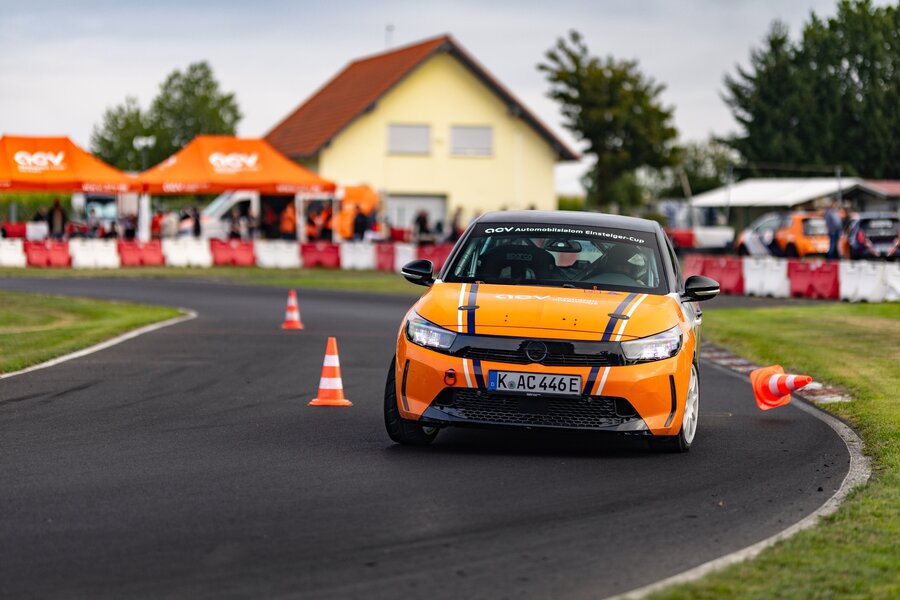 Ein orangefarbener Rennwagen rast auf einer Rennstrecke um eine Kurve und stößt dabei einen orangefarbenen Verkehrskegel um. Im Hintergrund sind Zelte und ein gelbes Haus zu sehen.