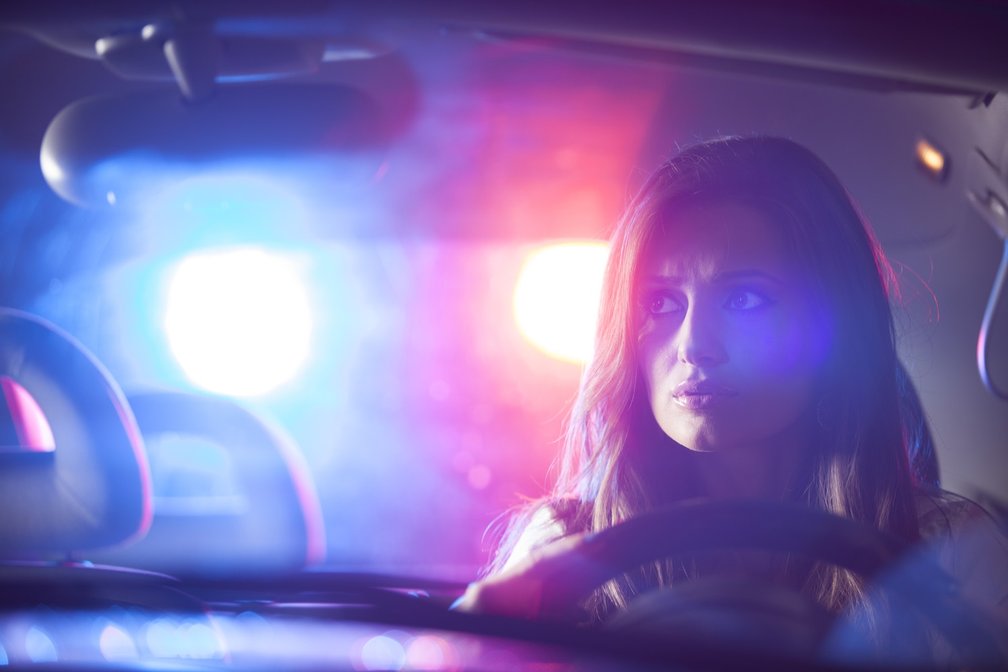 Junge Frau sitzt nachts am Steuer eines Autos, im Rückspiegel leuchten blau-rote Lichter eines Einsatzfahrzeugs–
