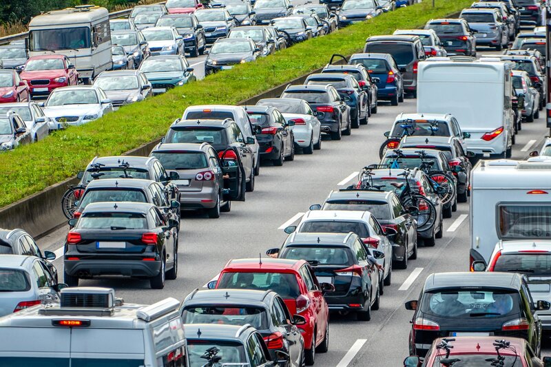 Starker Stau auf einer mehrspurigen Autobahn, auf der sich in beiden Fahrtrichtungen dicht aneinander gereihte Autos befinden, von denen einige Fahrräder transportieren. Ein grasbewachsener Mittelstreifen trennt die Gegenfahrbahnen voneinander.