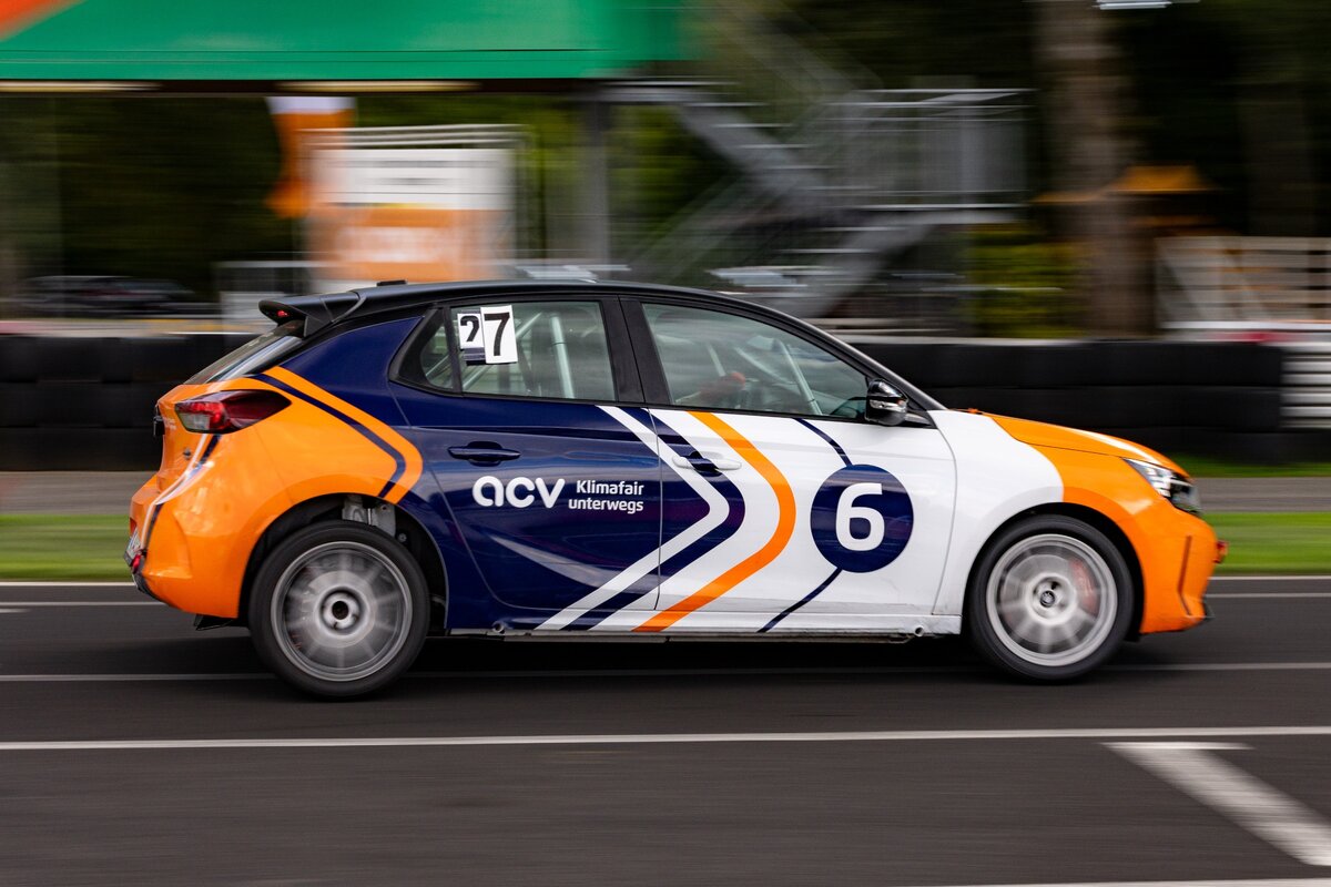 Ein Rennwagen mit orangefarbenen, weißen und blauen Streifen und der Startnummer 6 rast über eine Rennstrecke, im Hintergrund ist Bewegungsunschärfe zu sehen, und im Inneren ist der Fahrer zu erkennen.