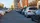 Eine belebte städtische Straße, gesäumt von parkenden Autos auf beiden Seiten. Im Hintergrund sind moderne Gebäude und eine teilweise bewölkte, sonnige Umgebung zu sehen.