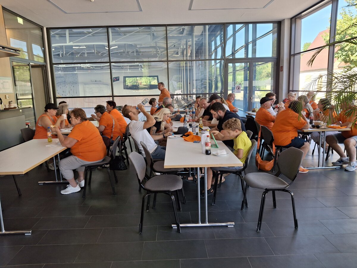 Eine Gruppe von Menschen, von denen viele orangefarbene Hemden tragen, sitzt an Tischen in einem hellen Raum mit großen Fenstern, isst, trinkt und unterhält sich. Es sind einige leere Stühle zu sehen. Es scheint sich um ein zwangloses Beisammensein oder eine Veranstaltung zu handeln.