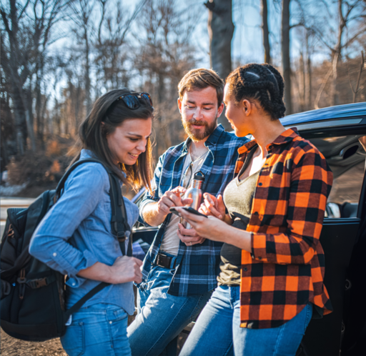 2 junge Frauen und ein junger Mann stehen angelehnt an ein Auto in einem Wald und zeigen auf ein Smartphone