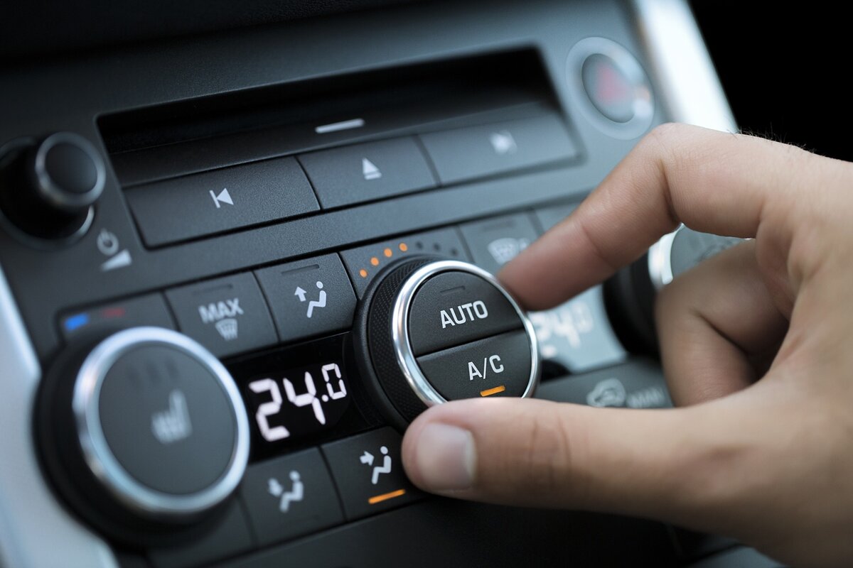 Eine Hand dreht den Drehregler „AUTO A/C“ am Bedienfeld der Klimaanlage eines Autos; auf dem digitalen Display ist die auf 24,0 °C eingestellte Temperatur zu sehen, und verschiedene Tasten für Luftstrom und Gebläsestufe sind sichtbar.