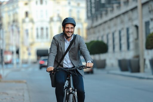 Ein lächelnder Mann mit Anzugjacke, Helm und Umhängetasche fährt mit dem Fahrrad auf einer Stadtstraße mit unscharfen Gebäuden im Hintergrund.