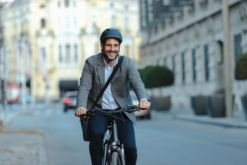 Ein lächelnder Mann mit Anzugjacke, Helm und Umhängetasche fährt mit dem Fahrrad auf einer Stadtstraße mit unscharfen Gebäuden im Hintergrund.