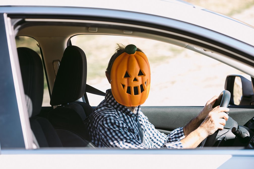 Eine Person sitzt am Steuer eines Autos und trägt eine große Kürbis-Maske mit geschnitztem Gesicht. Sie trägt ein kariertes Hemd und hält das Lenkrad mit beiden Händen.